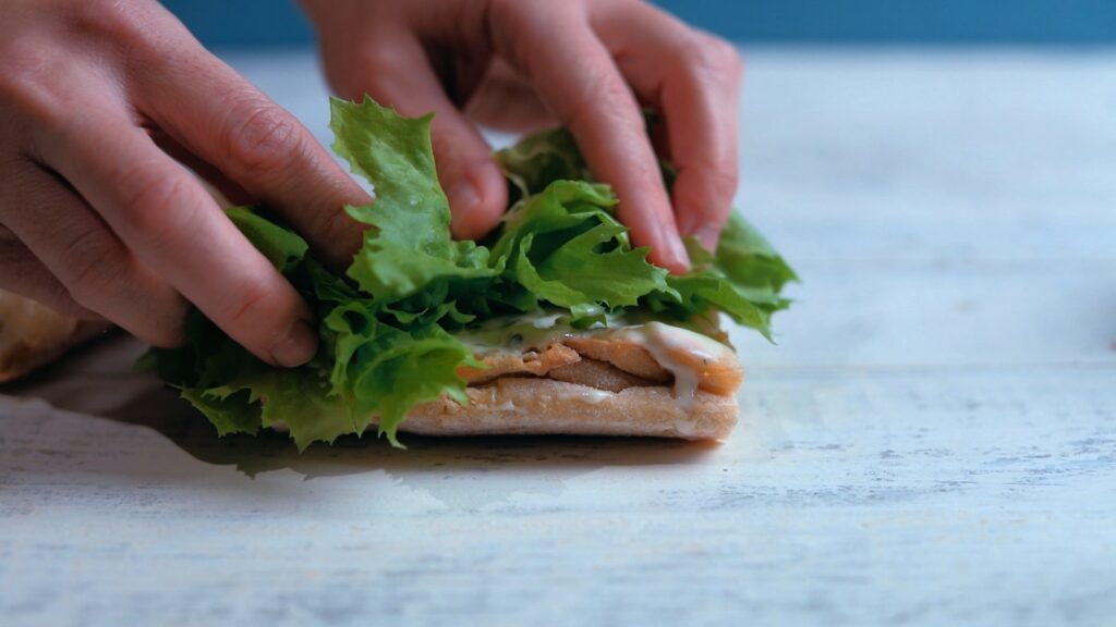 Close-up of hands delicately placing fresh salad greens on French bread with mayo dressing.