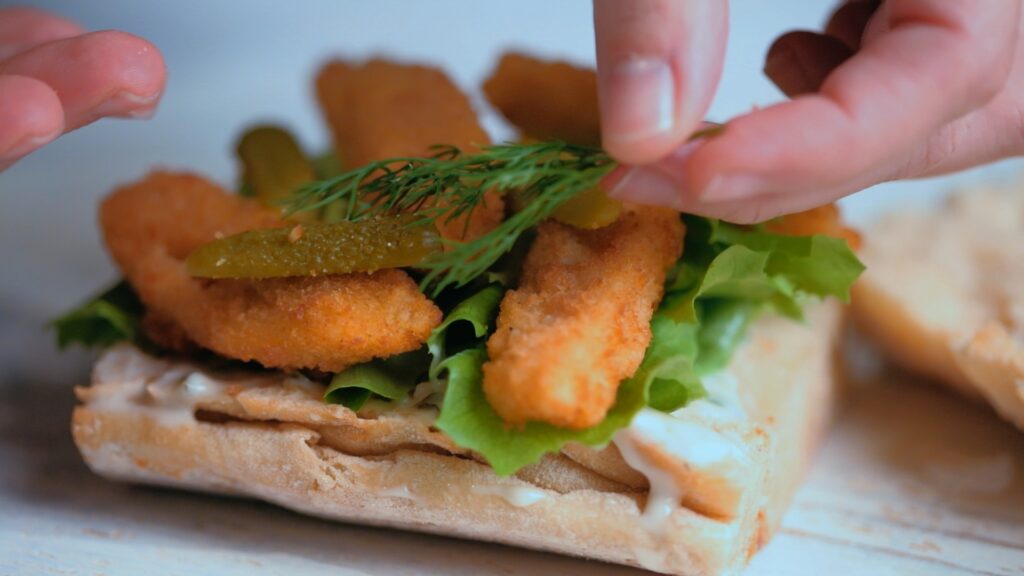 Extreme close-up of hands delicately placing garnish and baby pickles on a beautifully crafted gourmet sandwich. As a still from the video editing of this social media video.